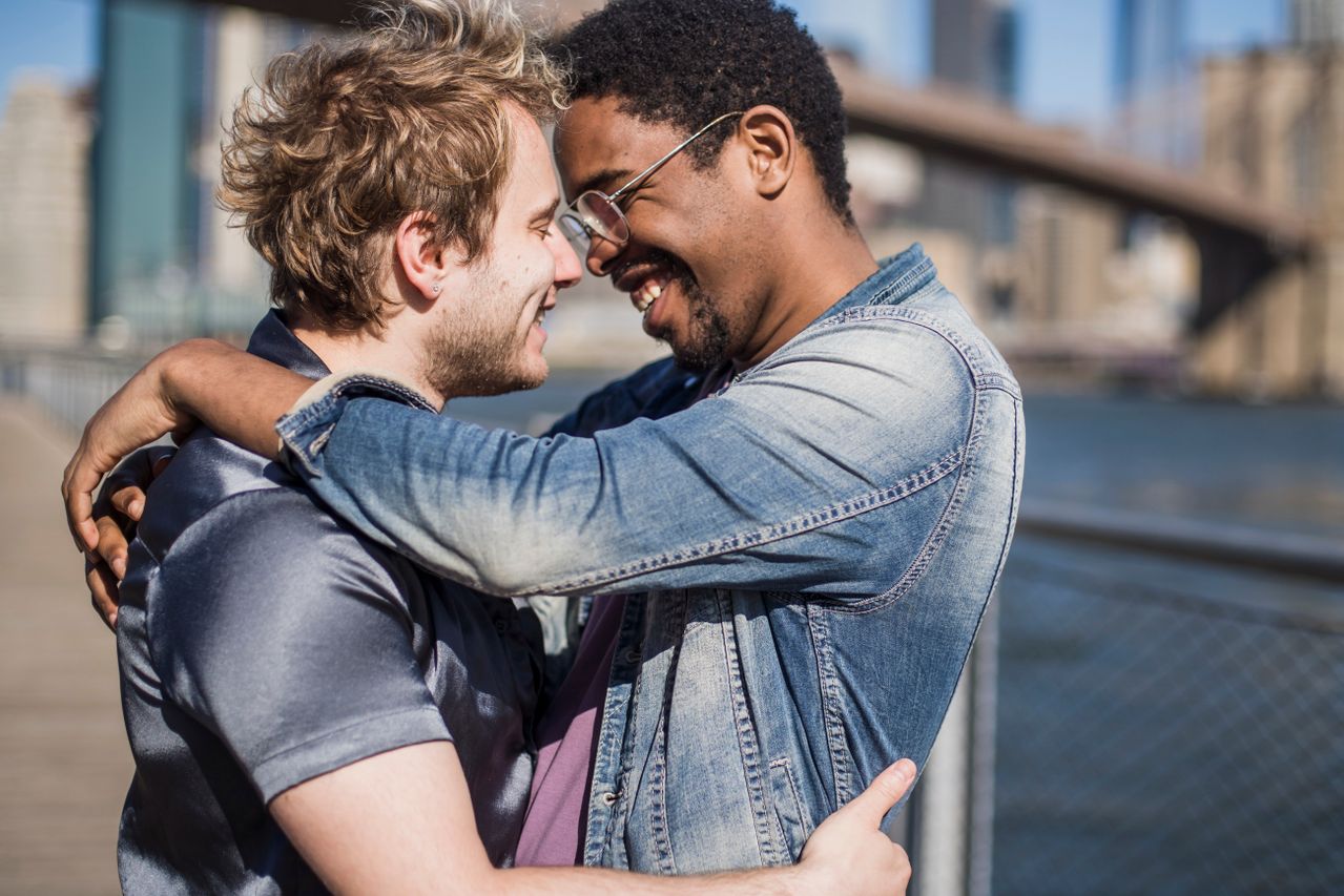 Two men hugging and laughing by the river with a bridge behind them
