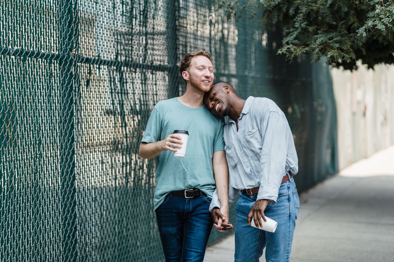 Man leaning on his partner's shoulder as they hold coffee by a green fence