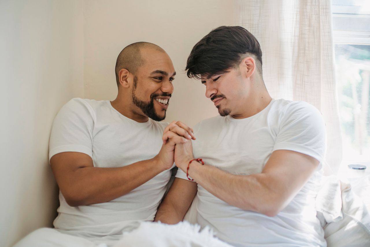 Two smiling men holding hands while sitting on a bed