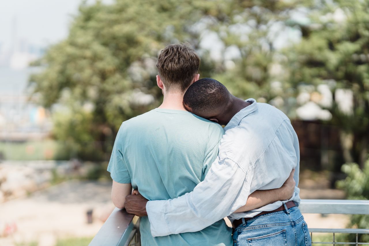 Two men hugging outdoors under green trees, seen from behind