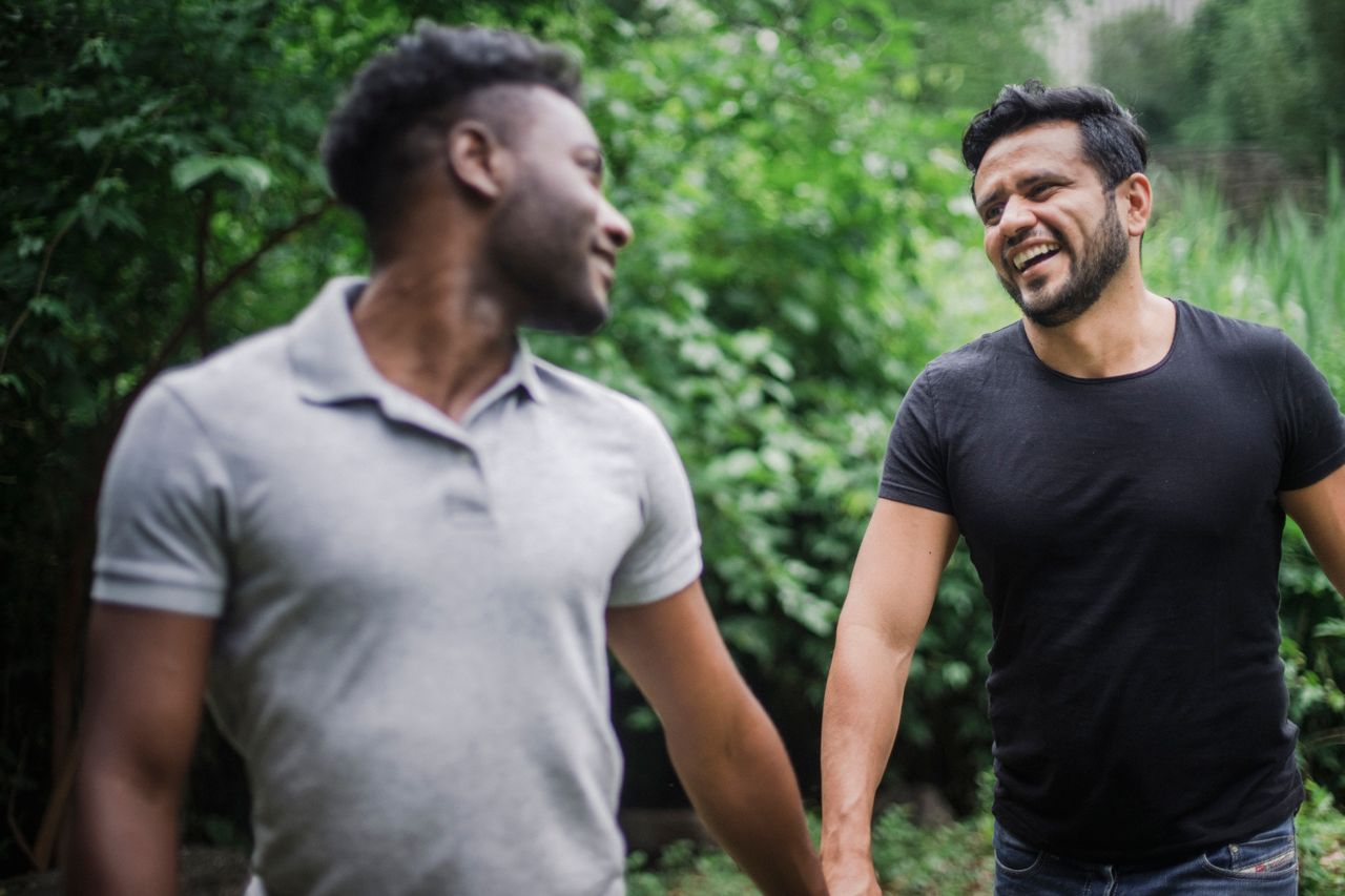 Two men laughing as they walk together past green trees