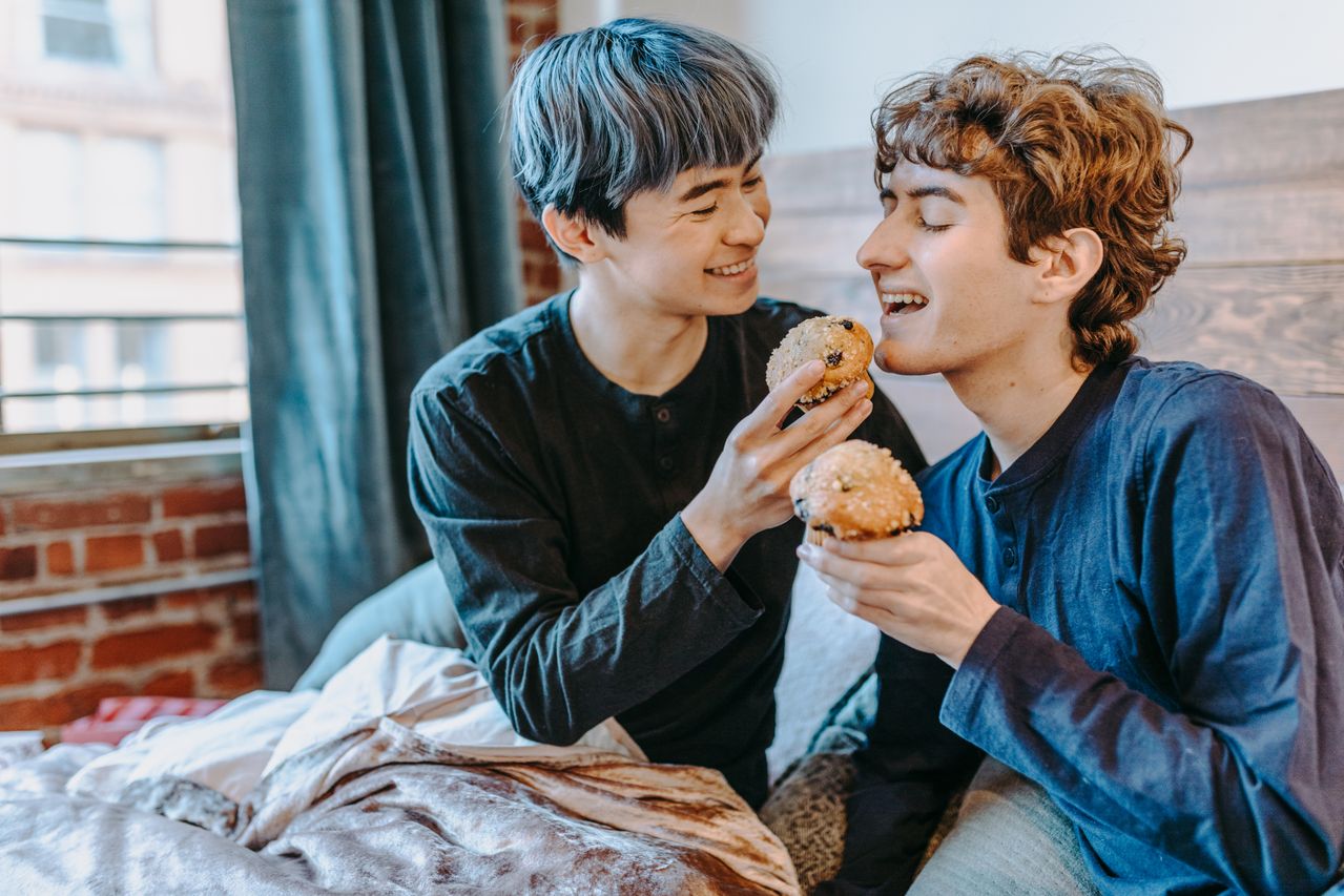 Two young men sharing pastries while sitting on a bed