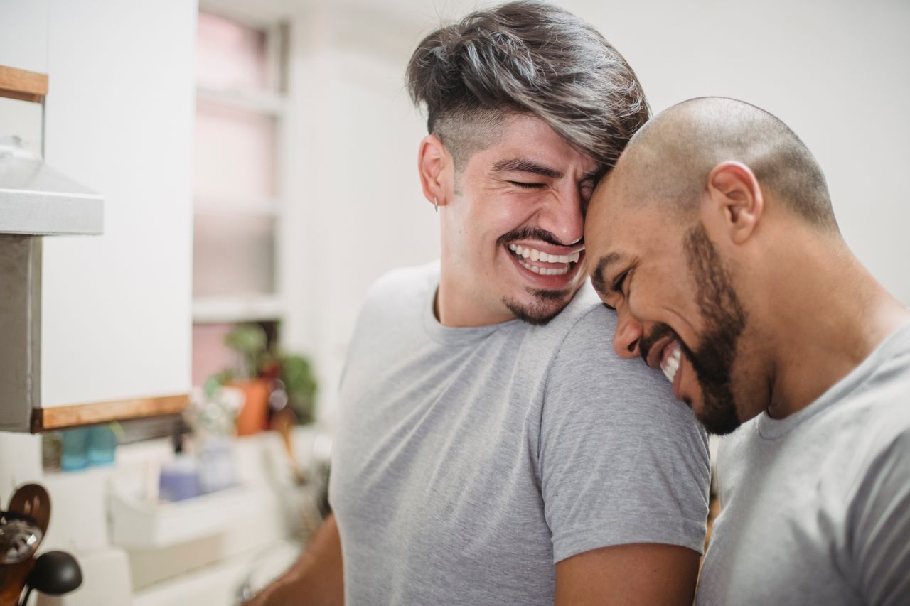 Two smiling men leaning their heads together in a kitchen