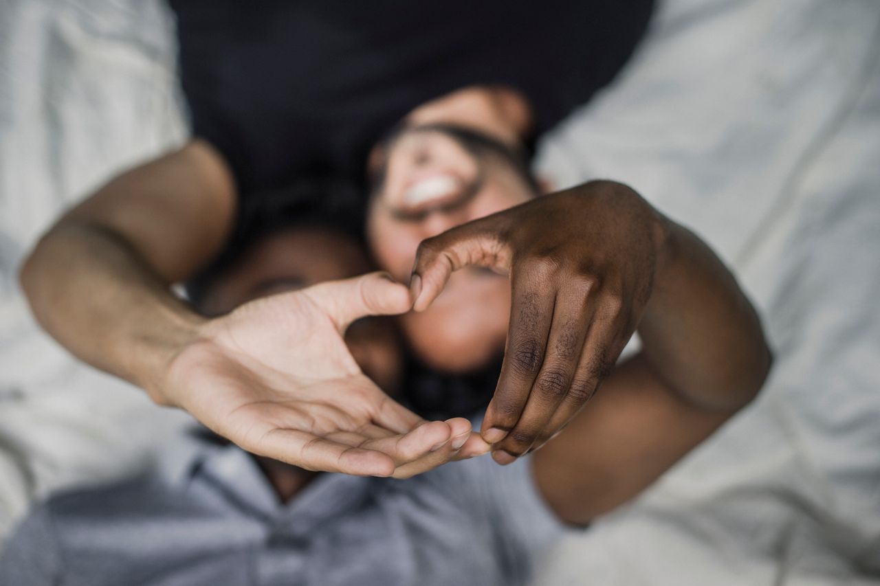 Two men lying in bed making a heart shape with their hands