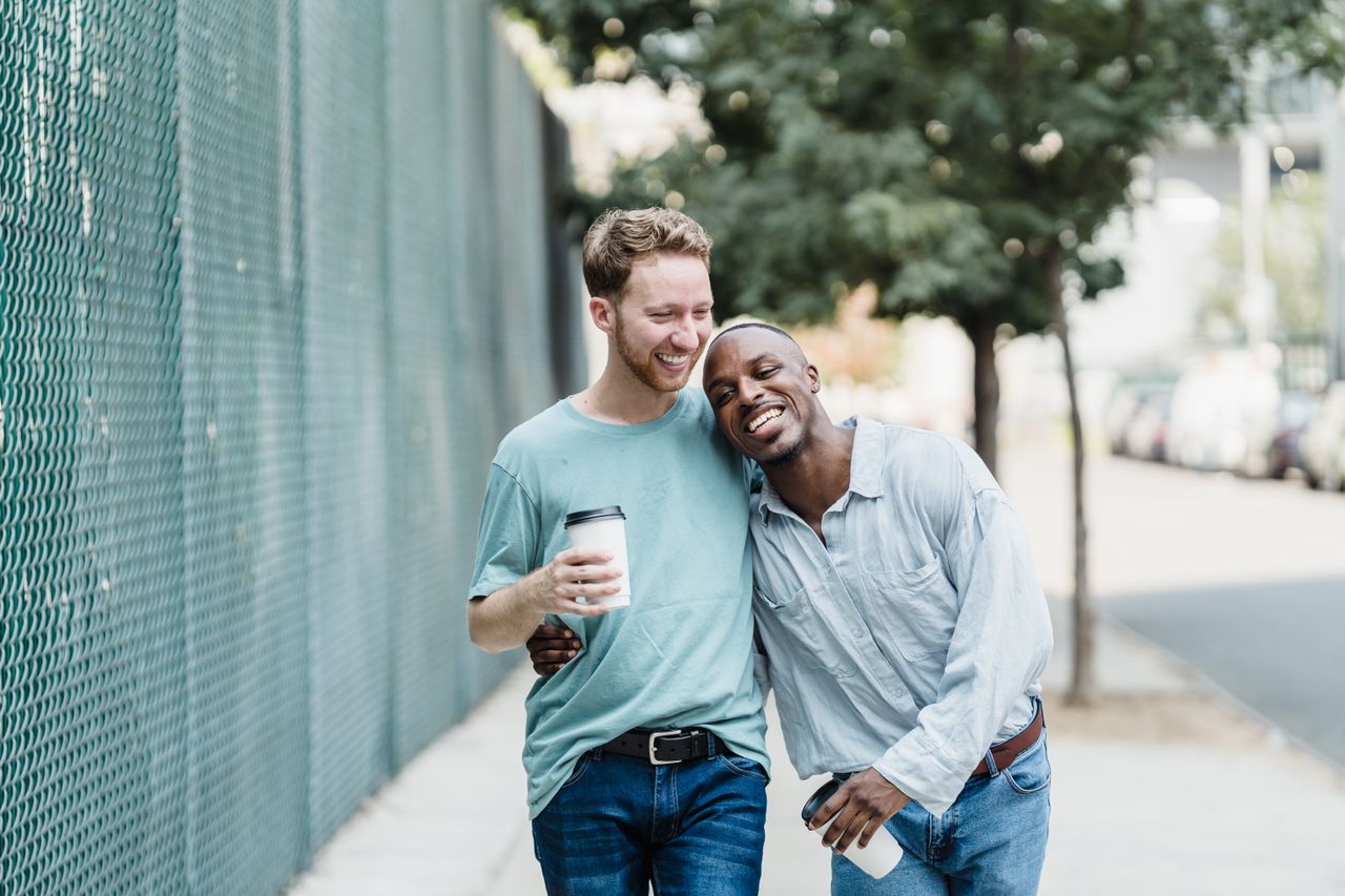 Two men walking with coffee, one leaning on the other's shoulder