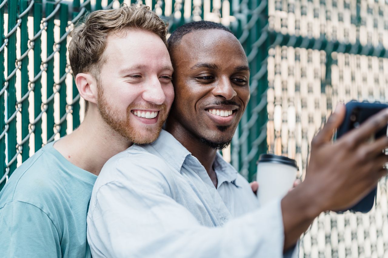 Two smiling men taking a selfie by a green fence