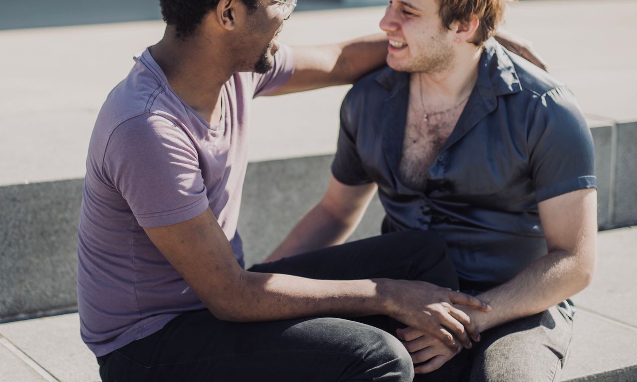 Two men sitting on steps outdoors, smiling and holding hands