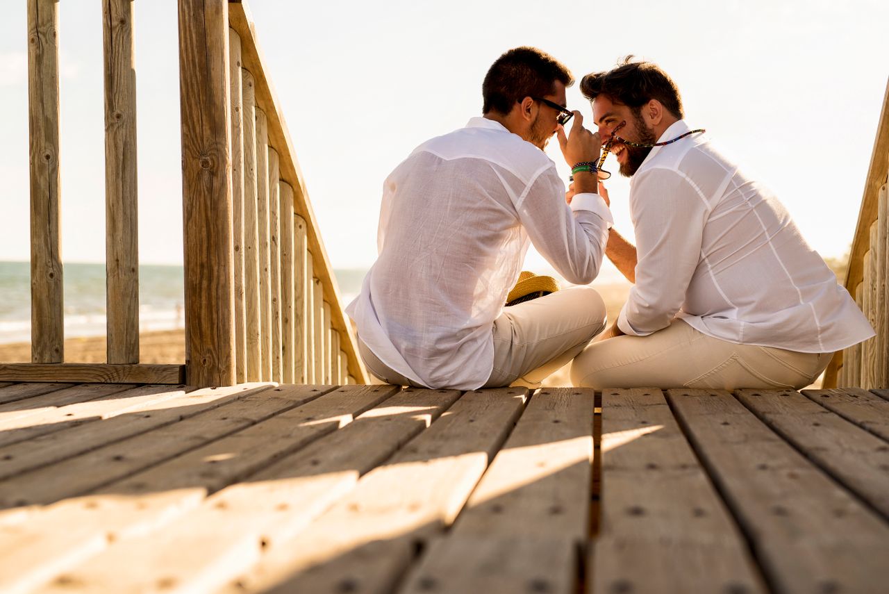 Two men in white shirts sitting close together on a wooden beach boardwalk