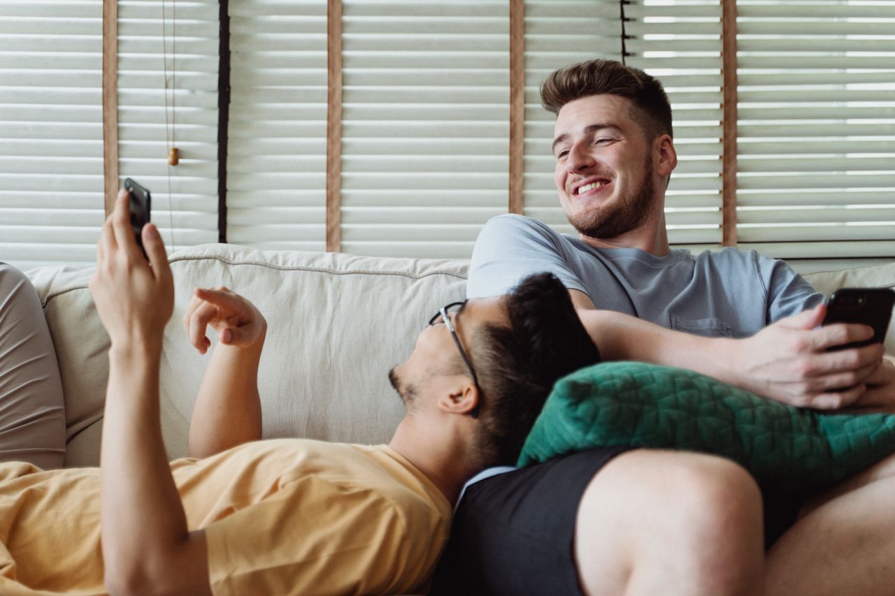 Two men relaxing on a sofa with their phones, one lying in the other's lap