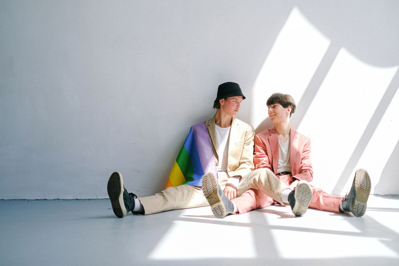 Two young men sitting on a sunlit floor with a rainbow flag