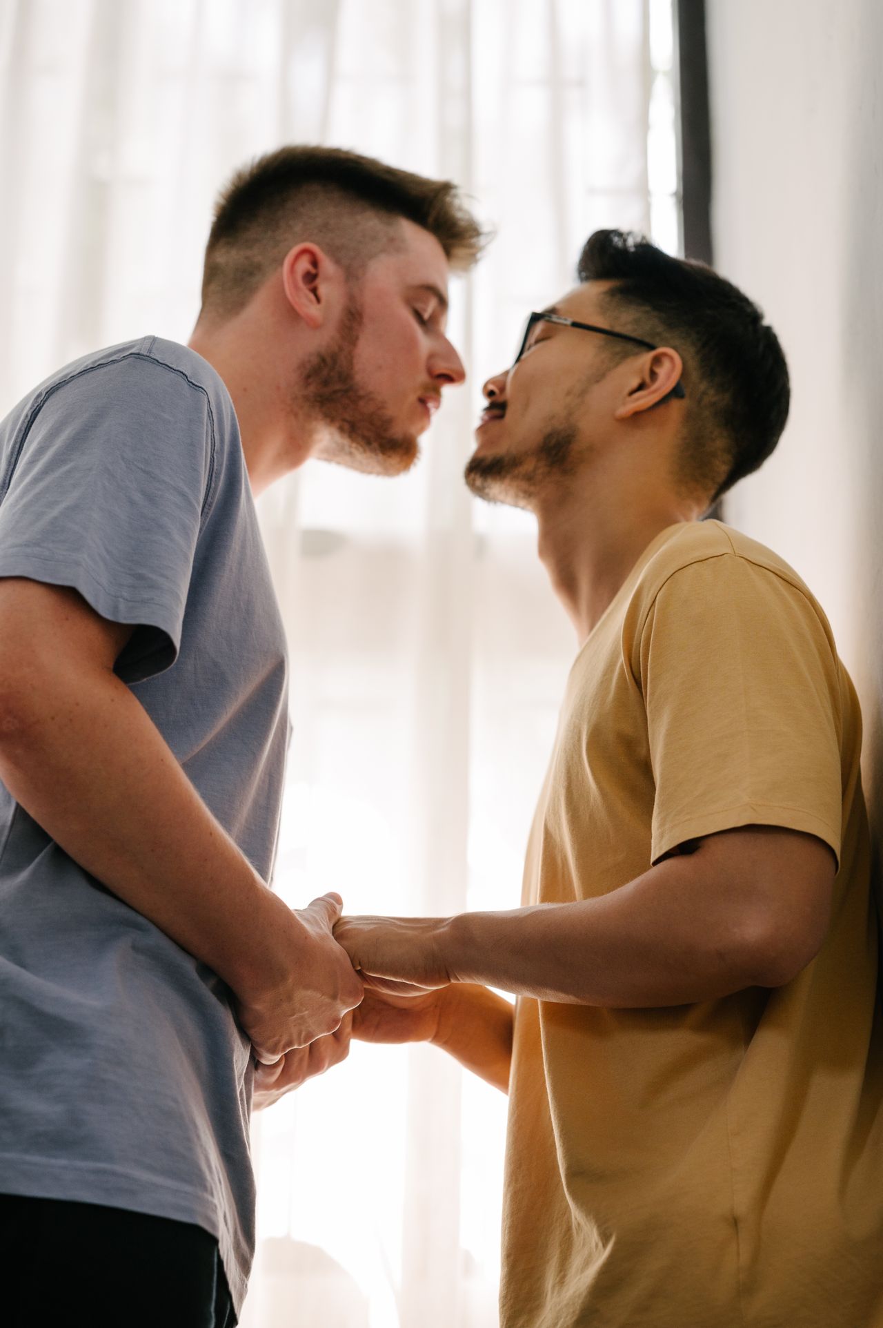 Two men holding hands and leaning in close by a bright window