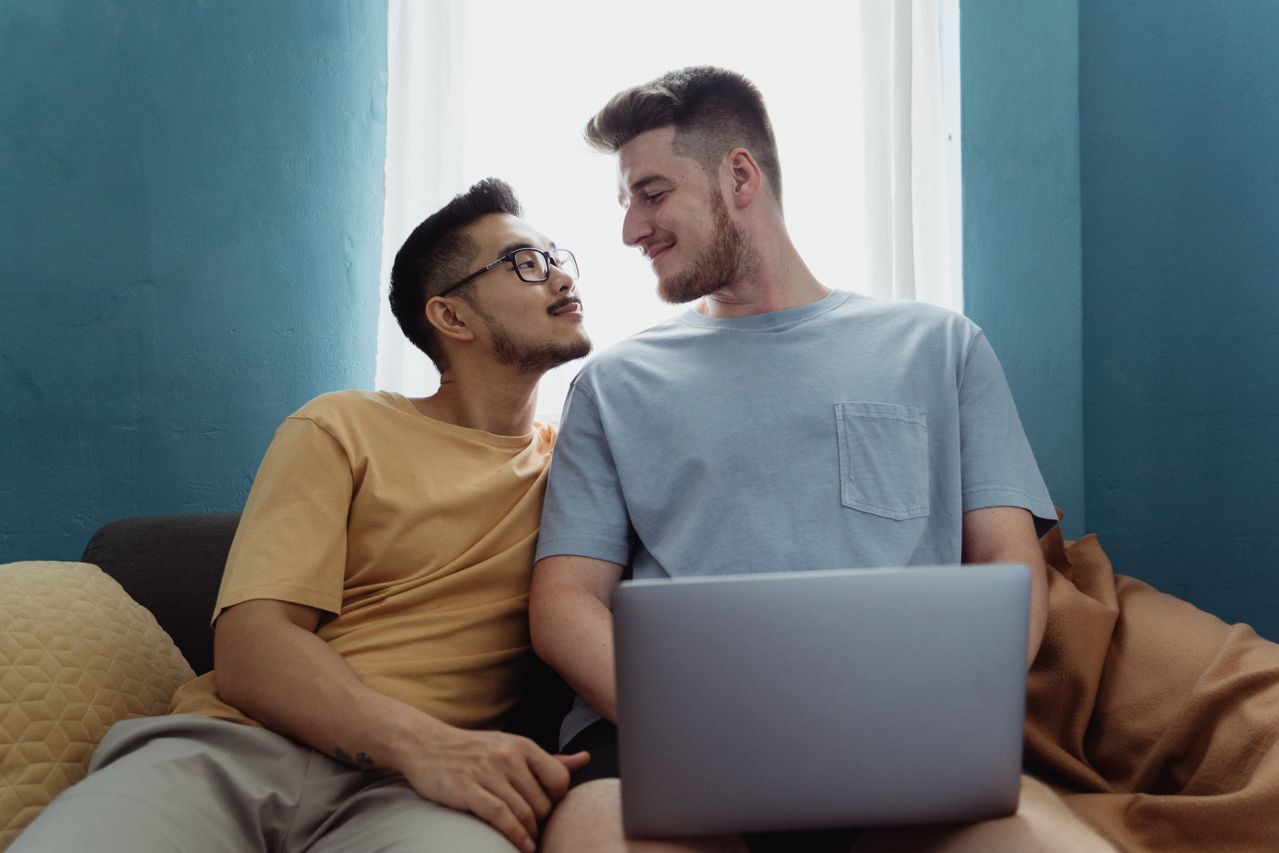 Two men smiling at each other on a sofa with a laptop