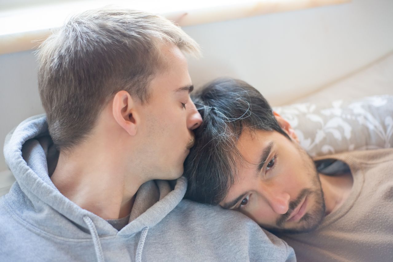 Man in a grey hoodie kissing his partner's head as they cuddle in bed